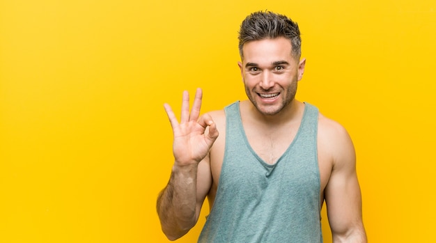 Hombre joven fitness contra un fondo amarillo guiña un ojo y tiene un gesto bien con la mano.