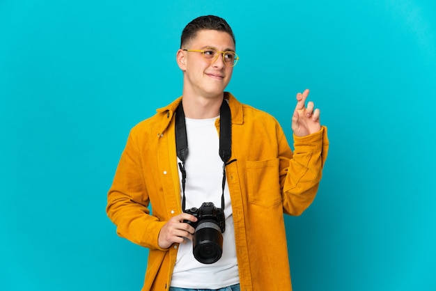 Hombre joven fotógrafo caucásico aislado en la pared azul con los dedos cruzando y deseando lo mejor