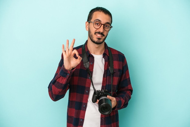 Hombre joven fotógrafo caucásico aislado sobre fondo azul alegre y confiado mostrando gesto ok.