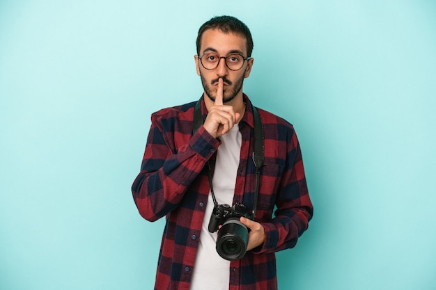 Hombre joven fotógrafo caucásico aislado sobre fondo azul manteniendo un secreto o pidiendo silencio.