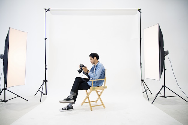 Hombre joven del fotógrafo que sienta la silla de madera y que comprueba el archivo en cámara en la escena blanca de centro en el estudio.