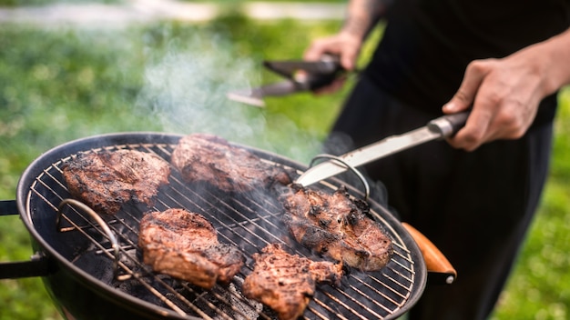 Hombre joven friendo carne a la parrilla. Verdor alrededor. Glamping