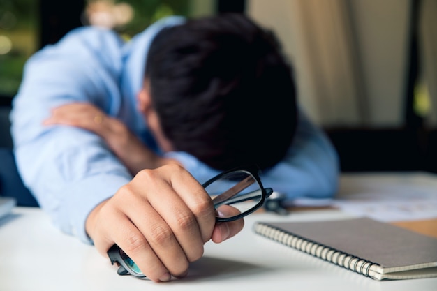 Hombre joven frustrado que parece exhausto mientras está sentado en su lugar de trabajo y llevando sus gafas en la mano.