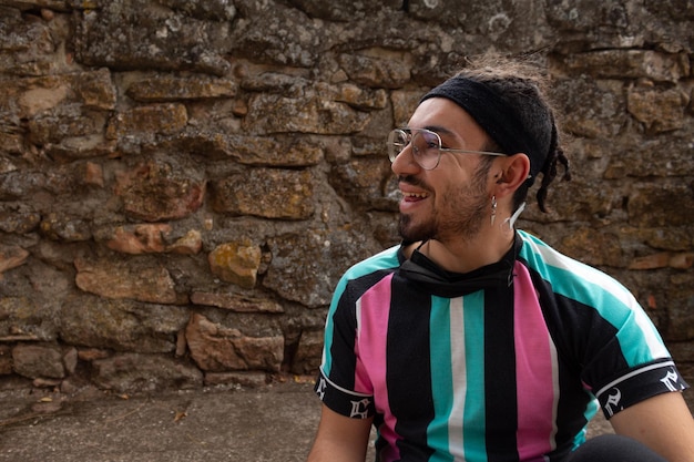 Foto hombre joven con gafas, barba y trenzas en el pelo y pendiente sonriendo feliz y mirando hacia el lado, fondo de pared de piedra