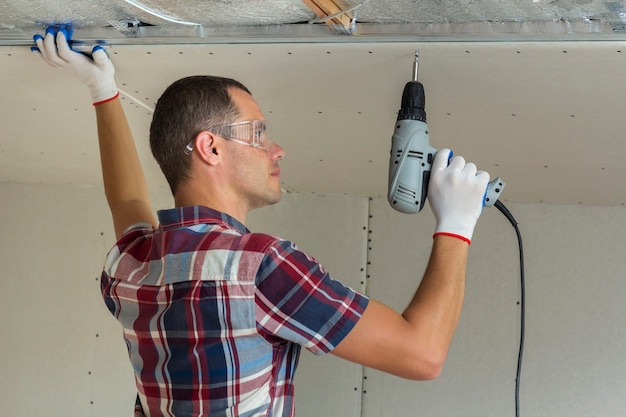 Hombre joven en gafas de fijación de paneles de yeso techo suspendido al marco de metal con un destornillador eléctrico en el techo.