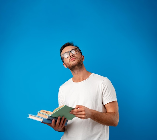 Hombre joven con gafas de pie y leyendo un libro contra el fondo azul.