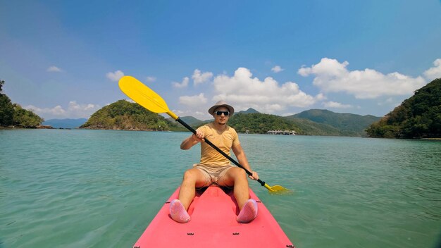 Hombre joven con gafas de sol y filas de sombreros en canoa de plástico rosa a lo largo del mar contra verdes islas montañosas con selvas salvajes Viajando a países tropicales El tipo fuerte navega en kayak en la vista frontal del océano