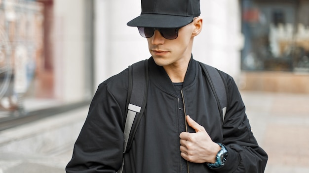 Hombre joven en gafas de sol de moda con una gorra negra y chaqueta con una bolsa en la ciudad