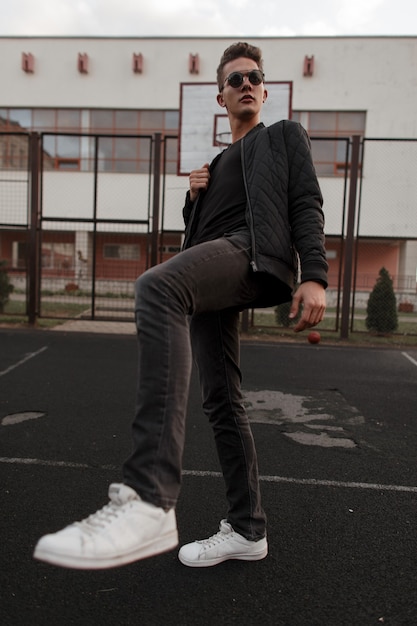 Hombre joven con gafas de sol en ropa negra de moda con una chaqueta negra y zapatillas blancas en el estadio en la calle