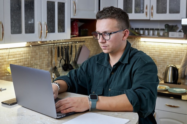 Hombre joven con gafas trabaja en su computadora portátil en autoaislamiento en casa. Concepto de tecnología, trabajo remoto y estilo de vida.