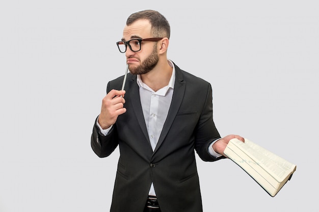 Hombre joven en gafas y traje de stands. Él sostiene la pluma cerca de los labios. Hay un gran libro en otra mano.