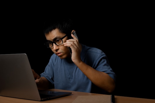 Hombre joven con gafas usando un teléfono inteligente y una computadora portátil en casa, trabaja desde el concepto de hogar sobre fondo negro.