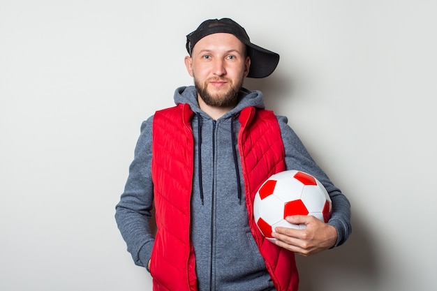 Hombre joven con una gorra, sudadera con capucha y chaleco casualmente vestidos sosteniendo un balón de fútbol contra una pared de luz