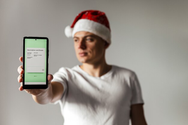 Hombre joven con gorro de Papá Noel tiene teléfono inteligente con certificado de vacunación covid electrónico