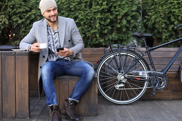 Hombre joven guapo con abrigo gris y sombrero con smartphone, descansando y sonriendo mientras está sentado cerca de su bicicleta al aire libre.