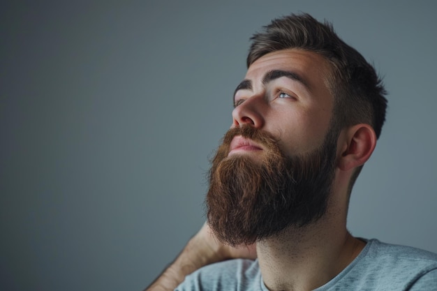 Hombre joven y guapo con barba está manteniendo la mano en la barba y mirando hacia otro lado en un fondo gris