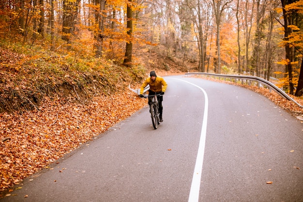 Hombre joven guapo en bicicleta por una carretera a través del bosque de otoño