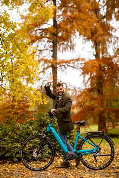 Hombre joven guapo con bicicleta eléctrica en el parque otoño