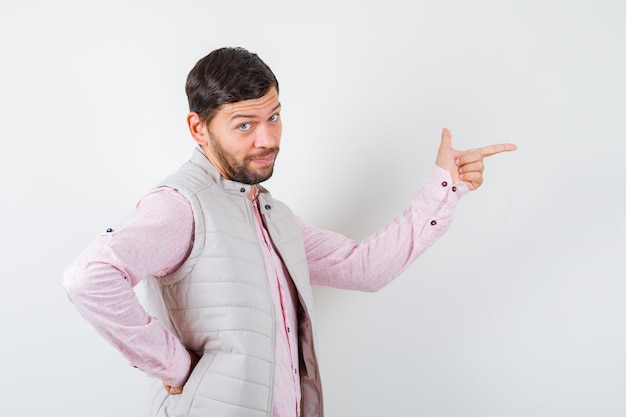 Hombre joven guapo en camisa, chaleco apuntando hacia la derecha, con la mano en la cintura y mirando confiado, vista frontal.