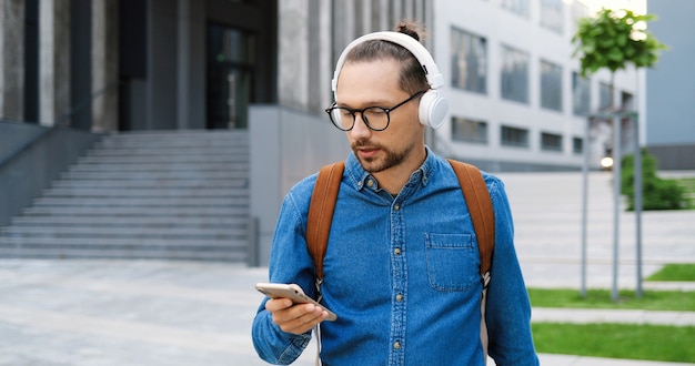 Hombre joven guapo caucásico en gafas tomando auriculares y escuchando música al aire libre en las calles de la ciudad. Un chico guapo con anteojos escucha una canción en la ciudad. Estudiante masculino con mochila.