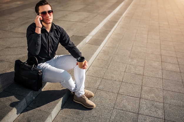 Hombre joven guapo en gafas hablar por teléfono. Él mira directamente a través de las gafas. Guy mantenga la taza de café en la mano. Se sienta afuera en los escalones.