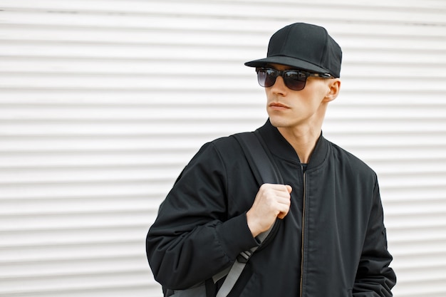 Hombre joven guapo con gafas de sol en una gorra de béisbol negra y chaqueta con una bolsa