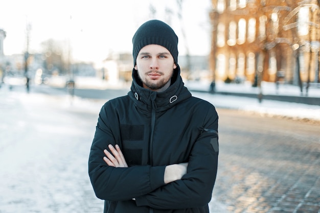 Hombre joven guapo con un gorro de punto y una chaqueta negra en un día de invierno al atardecer