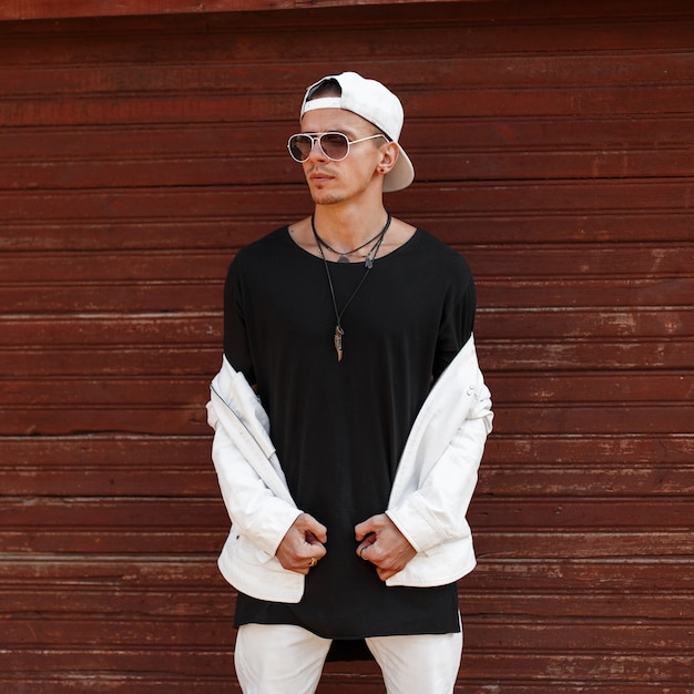 Hombre joven guapo hipster con una chaqueta de verano blanca de moda en una elegante camiseta negra con una gorra de béisbol de moda en gafas de sol oscuras cerca de una pared vintage de madera