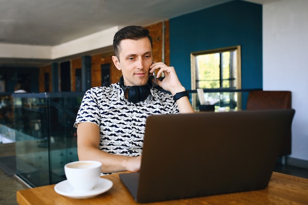 Hombre joven guapo hipster está usando laptop, hablando de teléfono móvil en la cafetería.