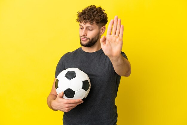 Hombre joven guapo jugador de fútbol aislado sobre fondo amarillo haciendo gesto de parada y decepcionado