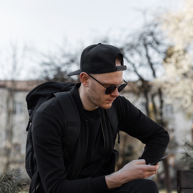 Hombre joven guapo de moda en elegantes gafas de sol con gorra en ropa negra con maletín se sienta y mira el teléfono móvil en la ciudad en un día soleado. Turista de chico atractivo descansando en la calle.