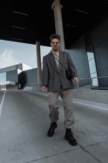 Hombre joven guapo de moda con gafas de sol vintage en un traje de moda con una chaqueta y una camisa está caminando en la ciudad cerca del edificio moderno