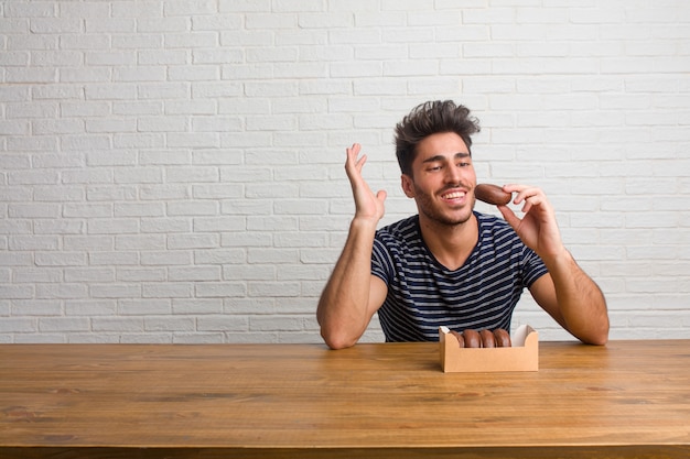 Un hombre joven, guapo y natural sentado en una mesa riendo y divirtiéndose, relajado y alegre, se siente seguro y exitoso. Comiendo donuts de chocolate.