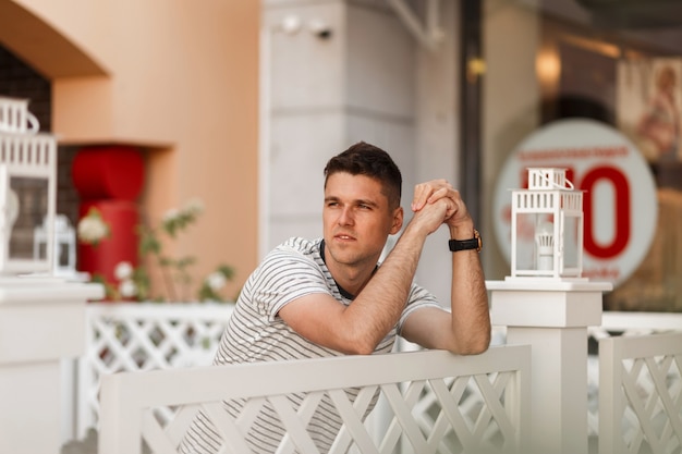 Hombre joven guapo con un peinado elegante en una camiseta de rayas de moda está de pie cerca de una valla de madera vintage