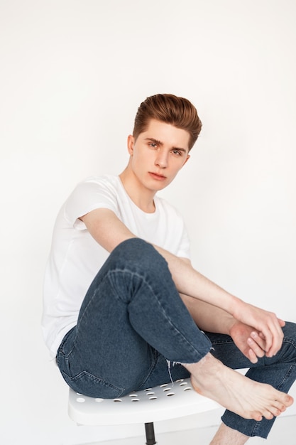 Hombre joven guapo con peinado de moda en camiseta blanca de moda en jeans con estilo se sienta en una silla sobre fondo blanco en estudio. Chico atractivo modelo posando junto a la pared vintage en la habitación.