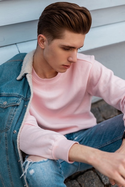 Hombre joven guapo con peinado de moda en ropa de jeans azul juvenil de moda de la nueva colección de verano descansa en la calle al aire libre.