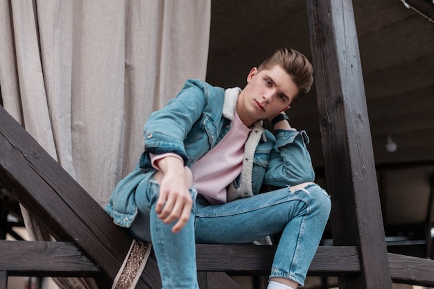 Hombre joven guapo con peinado de moda en ropa de jeans azul juvenil de moda de la nueva colección de verano descansa en la calle en la ciudad. Modelo de chico serio atractivo en ropa casual de mezclilla de moda al aire libre.