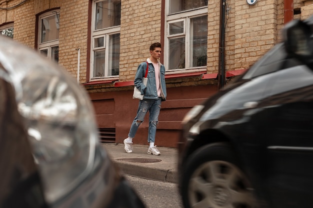 Hombre joven guapo con peinado en ropa de mezclilla casual azul de moda en zapatillas de deporte blancas de moda con bolso de tela vintage camina en la calle cerca de edificio de ladrillo y coches modernos. Chico atractivo a pie.