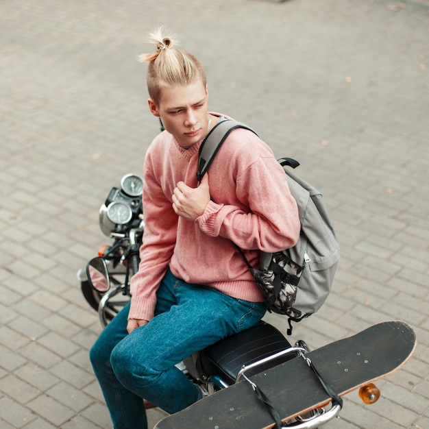 Hombre joven guapo con un peinado en un suéter rosa con una mochila y una patineta sentado en una motocicleta