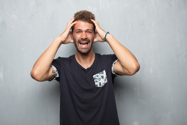 Un hombre joven y guapo que grita con una loca y sorprendida mirada de sorpresa, sosteniendo la cabeza con ambas manos.