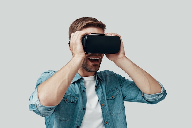 Hombre joven guapo con simulador de realidad virtual y sonriendo mientras está de pie contra el fondo gris