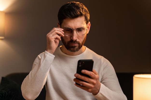 Un hombre joven y guapo sonriendo mientras se sienta con un smartphone y mira atentamente la pantalla. Foto de stock