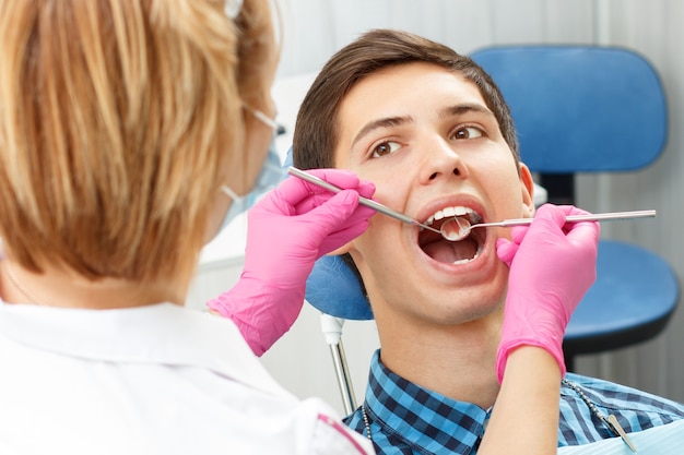 Hombre joven guapo de visita en el consultorio dental. Él está sentado en la silla y el dentista está examinando sus dientes con herramientas dentales. Concéntrese en la boca del paciente. Odontología