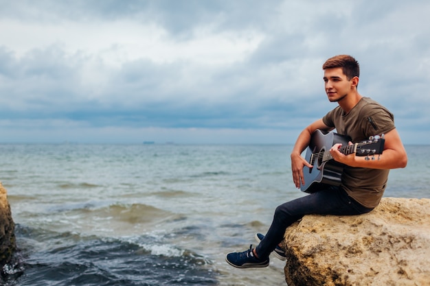 Hombre joven con guitarra acústica tocando en la playa rodeado de rocas en día lluvioso