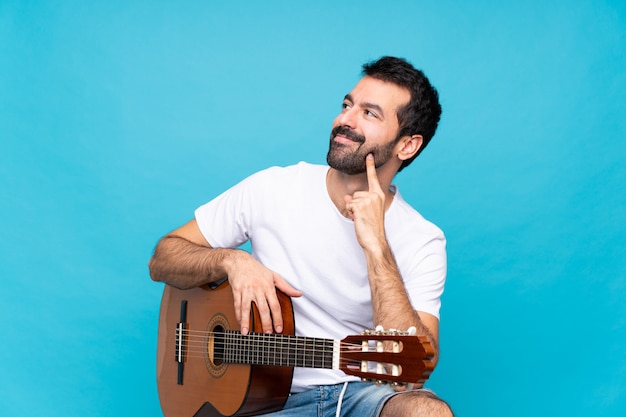 Hombre joven con guitarra sobre azul aislado pensando una idea mientras mira hacia arriba