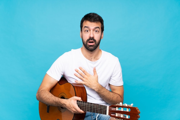 Hombre joven con guitarra sobre pared azul aislado sorprendido y conmocionado mientras mira a la derecha