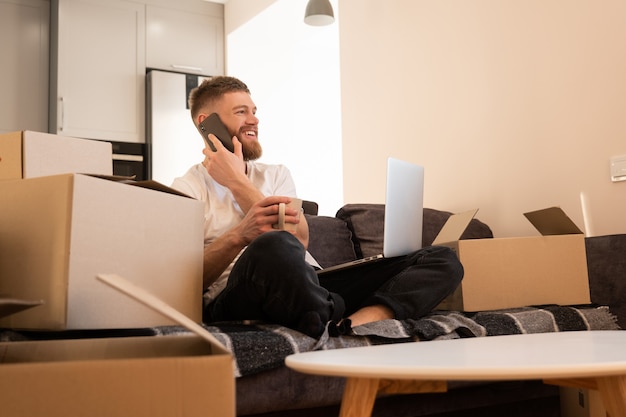 Hombre joven habla por teléfono móvil y bebe té o café. Chico europeo sonriente se sienta en el sofá y sostiene la computadora portátil. Cajas de cartón con cosas. Concepto de mudanza en piso nuevo. Interior del apartamento estudio