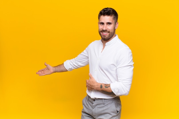 Hombre joven handosme sintiéndose feliz y alegre, sonriendo y dándole la bienvenida, invitándolo con un gesto amistoso contra la pared de color