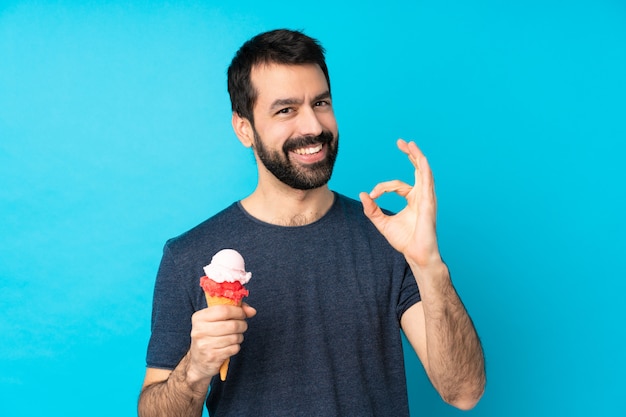 Hombre joven con un helado de cucurucho sobre la pared azul aislada que muestra la muestra aceptable con los dedos