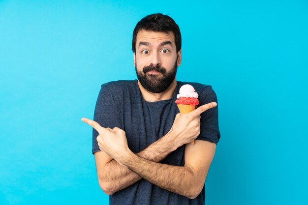 Hombre joven con un helado de cucurucho sobre pared azul aislado apuntando a los laterales que tienen dudas